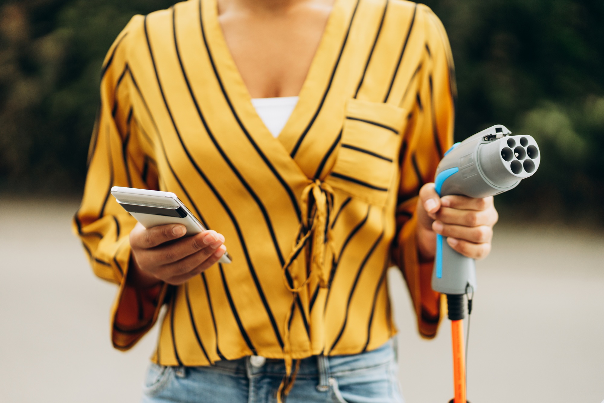 Close up of woman holding plug of the charger for an electric car and mobile phone outdoors